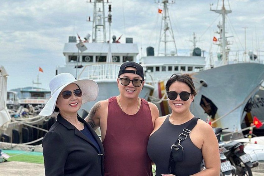 Three people standing in front of a large ship with a dock and other boats in the background. 2
