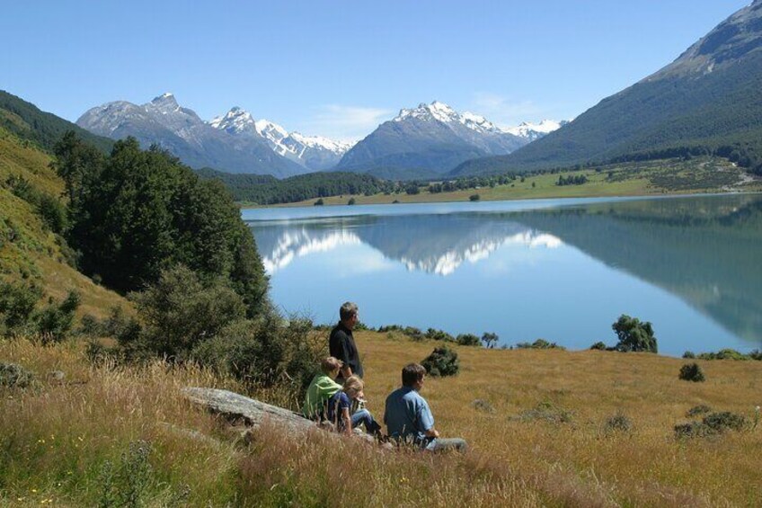 A lake with mountains in the background, a person sitting on the grass, and another person standing nearby. 4