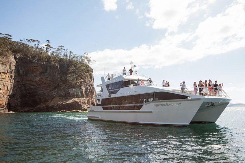 A white boat named 'Peppermint Bay Cruises' with passengers on deck, near a rocky cliff and calm waters. 1