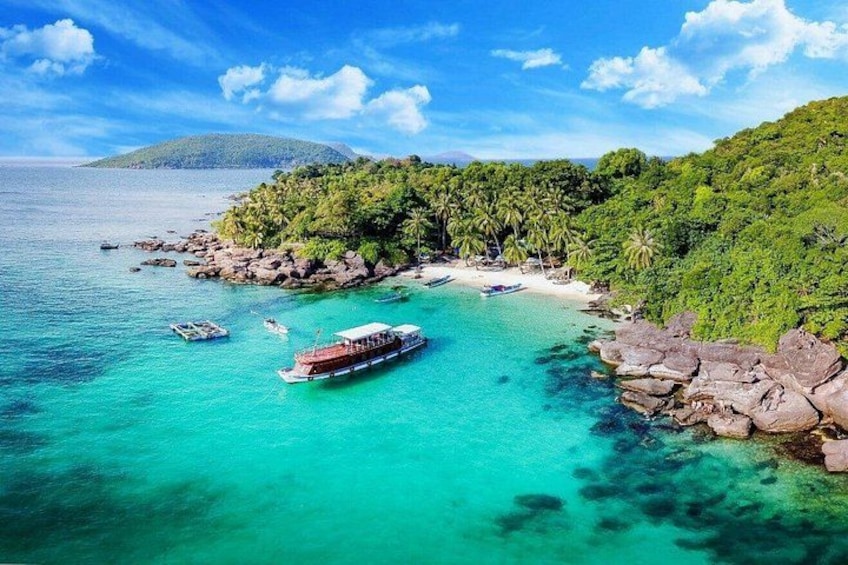 A tropical beach with clear turquoise waters, a large boat, and a rocky shoreline with dense greenery. 2