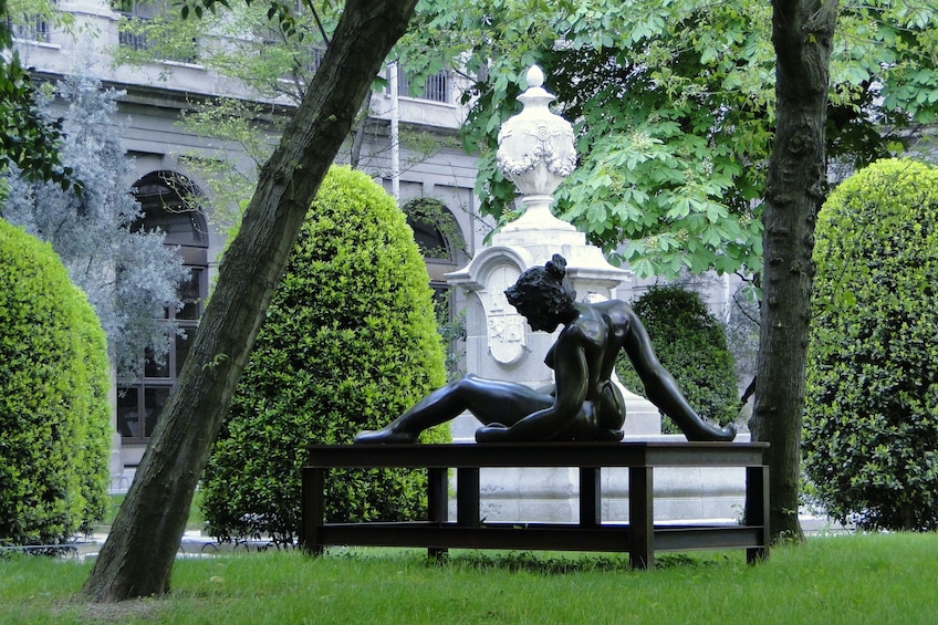 A sculpture of a reclining figure on a bench in a park with trimmed hedges and trees. 5