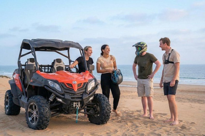 Escena de playa con un buggy aparcado en la arena, gente reunida y el océano al fondo. 1