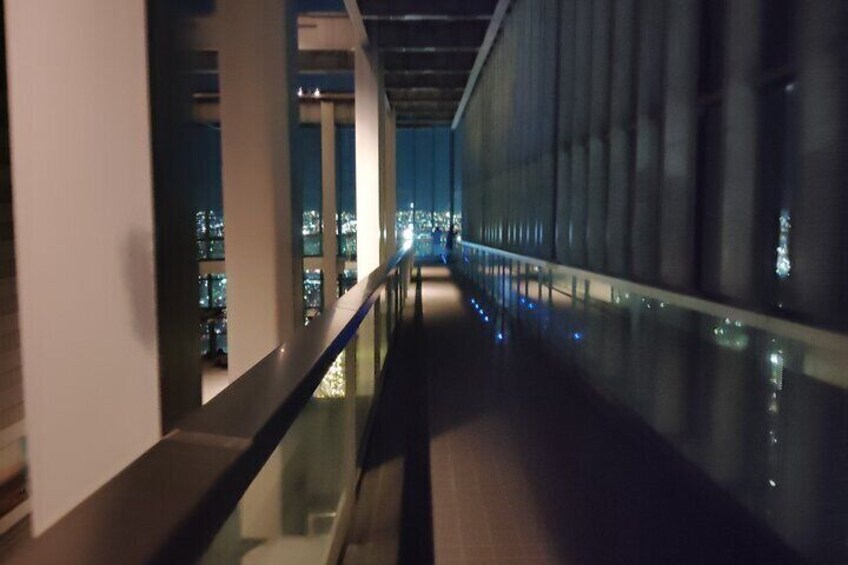 Vista nocturna de un pasillo con paredes de vidrio y barandilla, que conduce a un paisaje urbano con edificios iluminados. 4