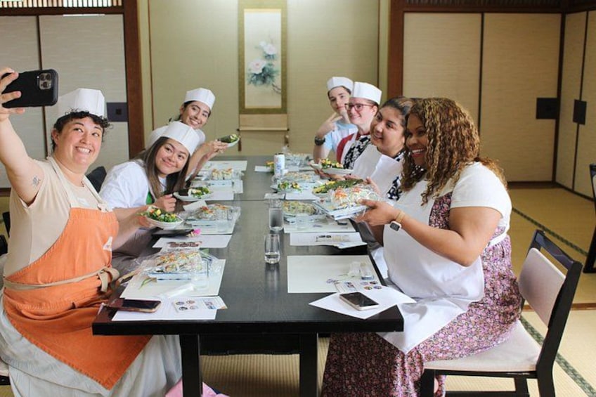 Un grupo de personas con gorros y delantales de chef, sentadas alrededor de una mesa con comida y papeles, en una sala de decoración tradicional. 5