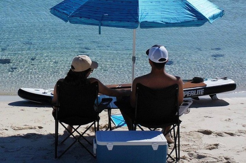 Two people sitting under a blue umbrella on a sandy beach, with a kayak and a cooler nearby. 2