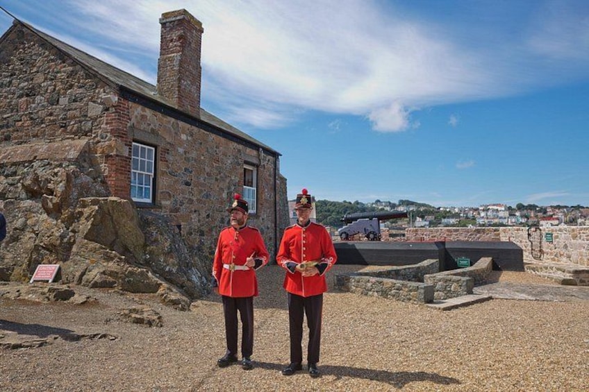 Two individuals in red uniforms standing in front of a stone building with a cannon in the background. 3