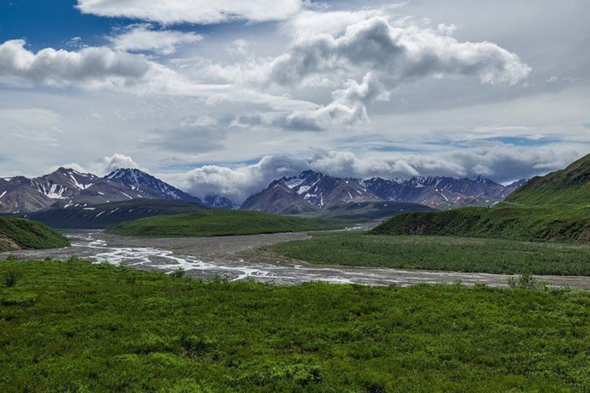 A mountainous landscape with a river, green vegetation, and snow-capped peaks under a cloudy sky. 3