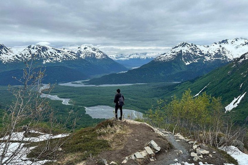 A person standing on a mountain ridge overlooking a valley with a river and snow-capped mountains. 5