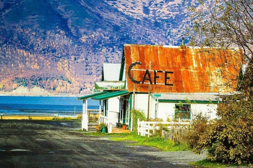 A rustic cafe with a large 'CAFE' sign, a covered outdoor seating area, and a view of a lake and mountains in the background. 3