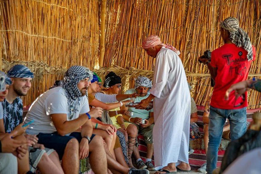A group of people in traditional attire seated and standing in a thatched-roof structure. 5