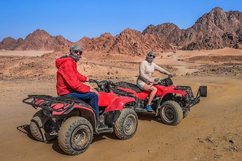 Two people on red ATVs in a desert landscape with mountains in the background. 3