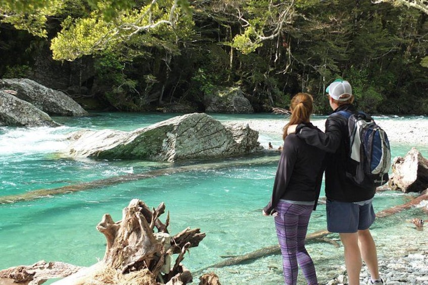 Two people standing by a river with clear water, surrounded by rocks and trees. 5