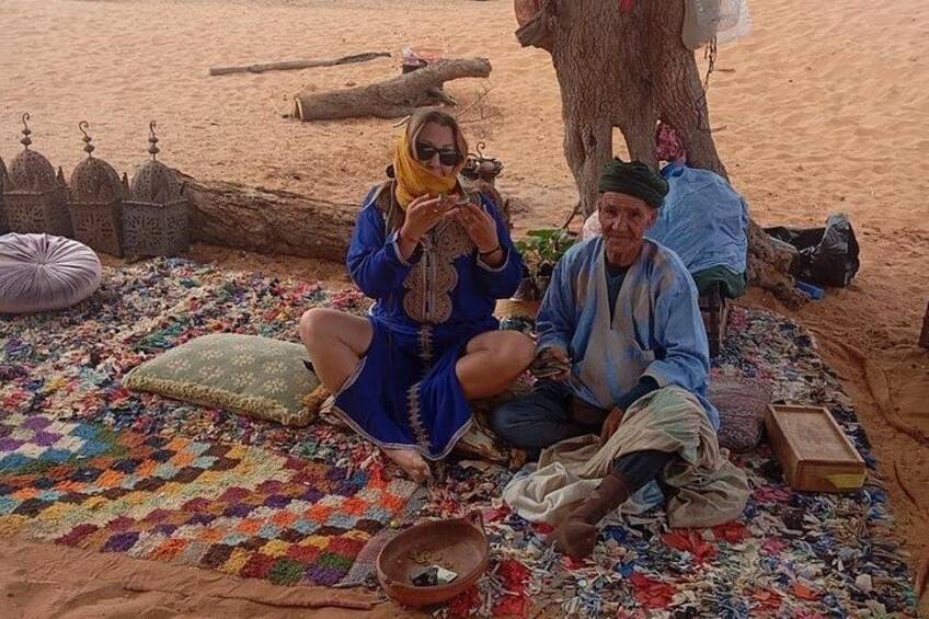 Two people sitting on a colorful blanket in a desert setting with a tree and traditional architecture in the background. 5