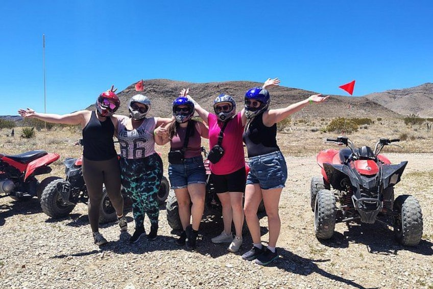 Four people in helmets and protective gear standing with arms outstretched in a desert landscape with ATVs in the background. 5