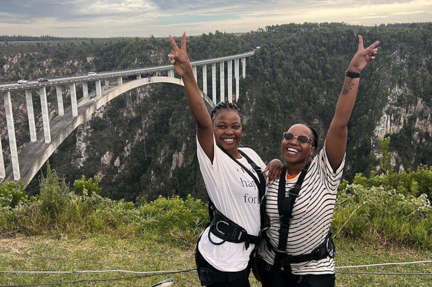Two people standing in front of a bridge with a mountainous landscape in the background. 5