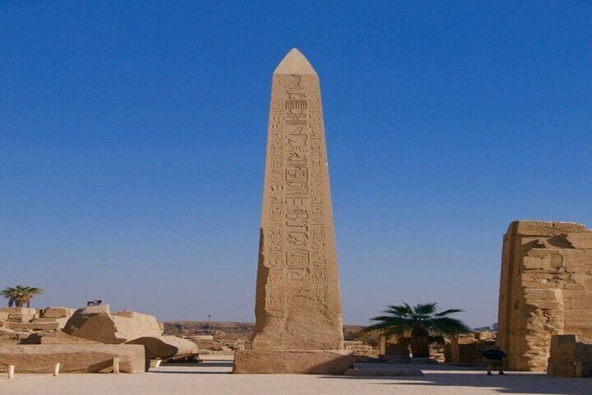 Ein hoher, antiker ägyptischer Obelisk mit Hieroglyphen, umgeben von Ruinen und Palmen. 3