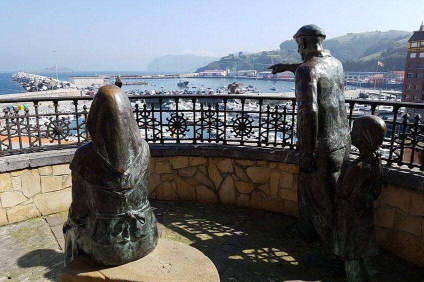 Une statue représentant une femme et un homme, placée sur un socle en pierre surplombant un port où se trouvent des bateaux, avec une côte lointaine en arrière-plan. 2