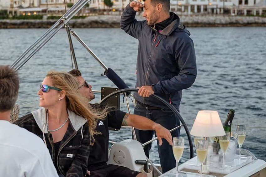 A group of people on a boat with a table set for a meal, glasses of wine, and a person standing at the helm. 2