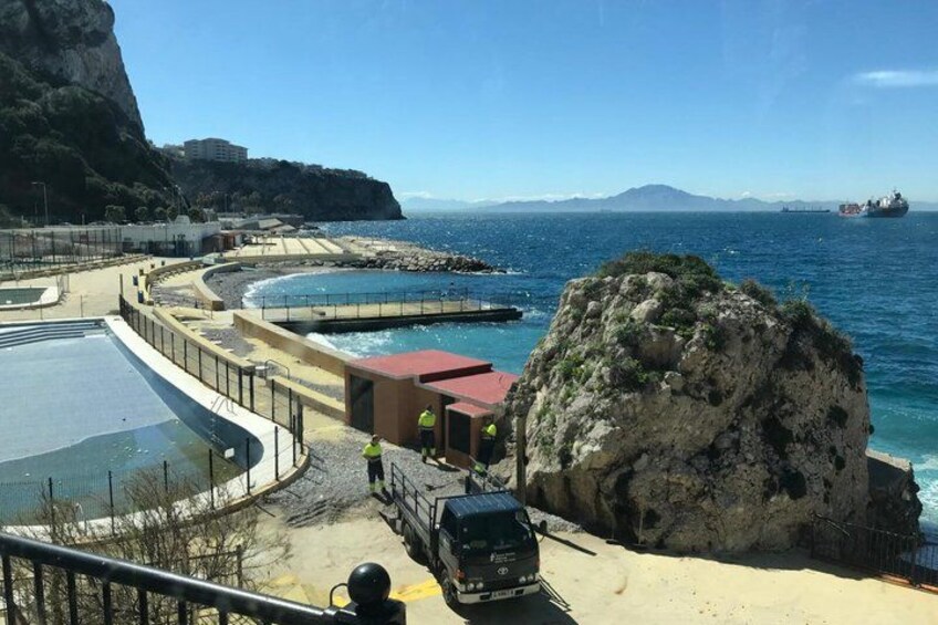 Ett kustområde med en stor klippformation, en swimmingpool och ett fartyg i bakgrunden. 2