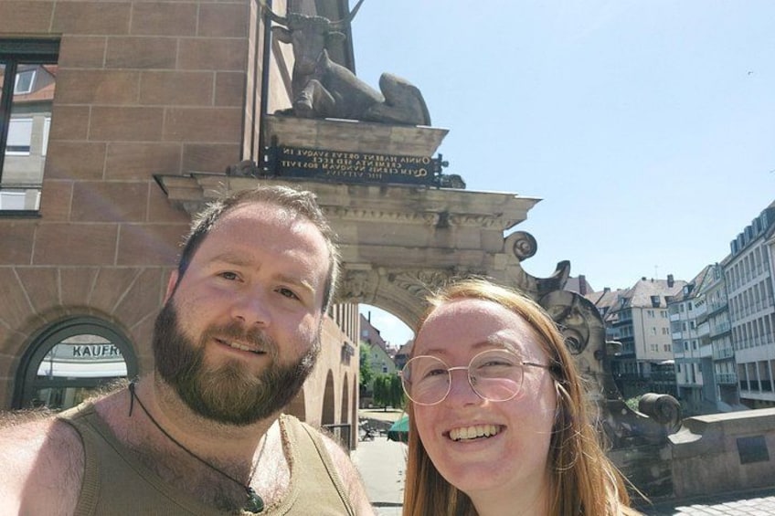 A man and a woman standing in front of a stone archway with inscriptions, with buildings in the background. 4