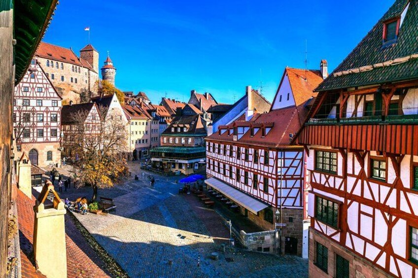 A cobblestone street lined with traditional half-timbered buildings, a castle tower, and a clear blue sky. 1