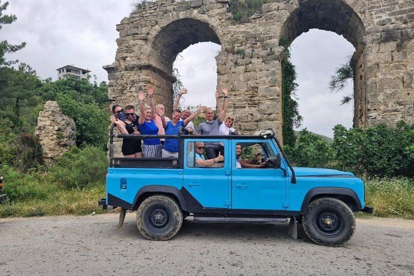 A blue off-road vehicle with passengers, parked in front of ancient stone arches. 1