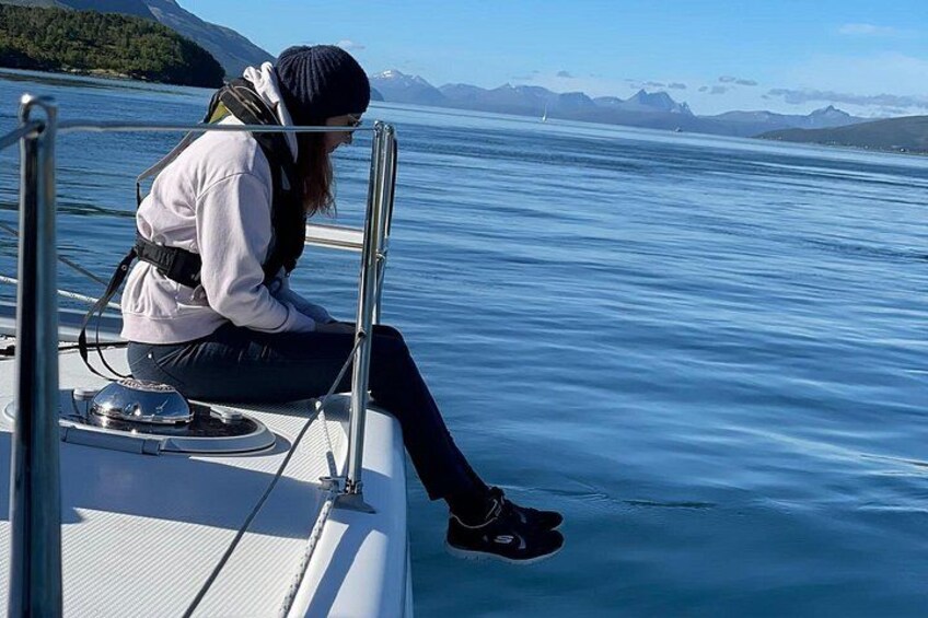 Eine Person sitzt in einem Boot, trägt eine Mütze und eine Jacke, im Hintergrund ruhiges Meer und Berge. 2