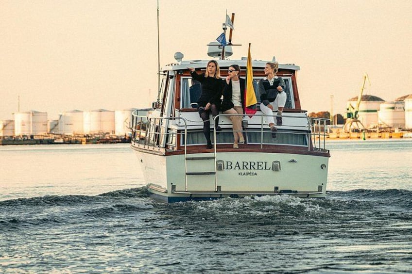 En båt vid namn ”Barrel” med tre personer ombord, som seglar på vattnet med industriella byggnader i bakgrunden. 2