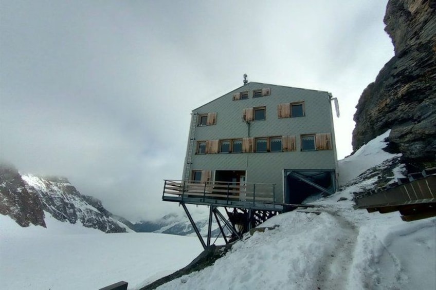 Un rifugio di montagna con una scala in metallo, montagne innevate e un cielo nuvoloso. 3