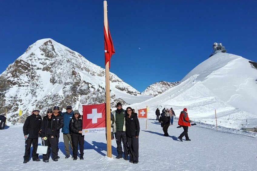 Eine Gruppe von Personen steht in einer verschneiten Landschaft, im Hintergrund ist ein Berg zu sehen und ein Fahnenmast mit einer Flagge. 2