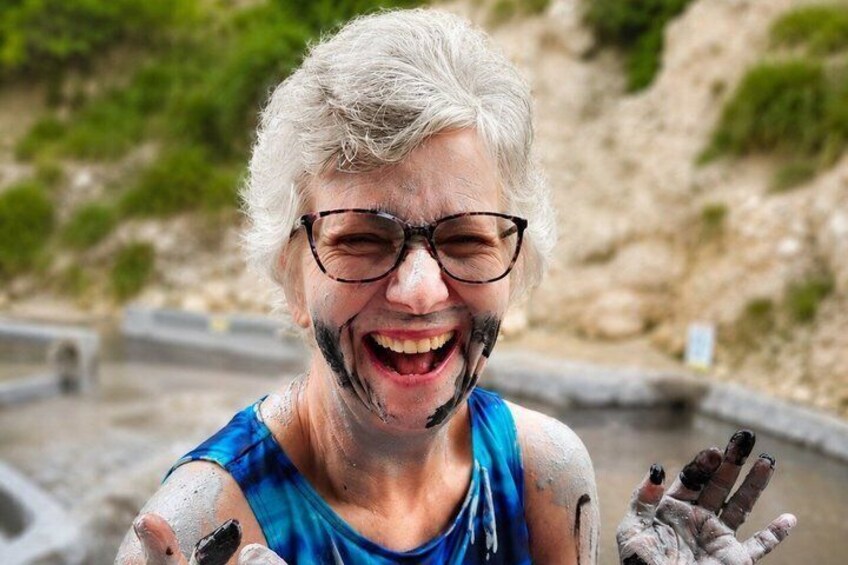 A person covered in mud, wearing a blue tank top, with a rocky landscape in the background. 1