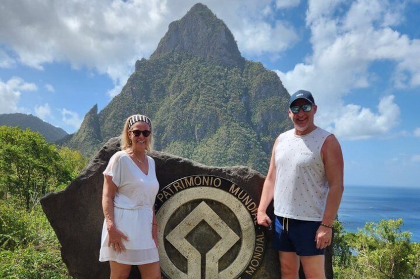 Two people standing in front of a sign with 'Patrimonio Mundo' written on it, with a mountainous landscape and ocean in the background. 2