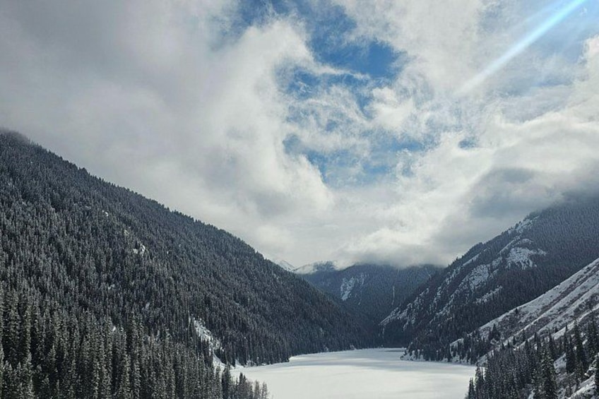A snow-covered lake surrounded by dense forests and mountains under a cloudy sky. 1