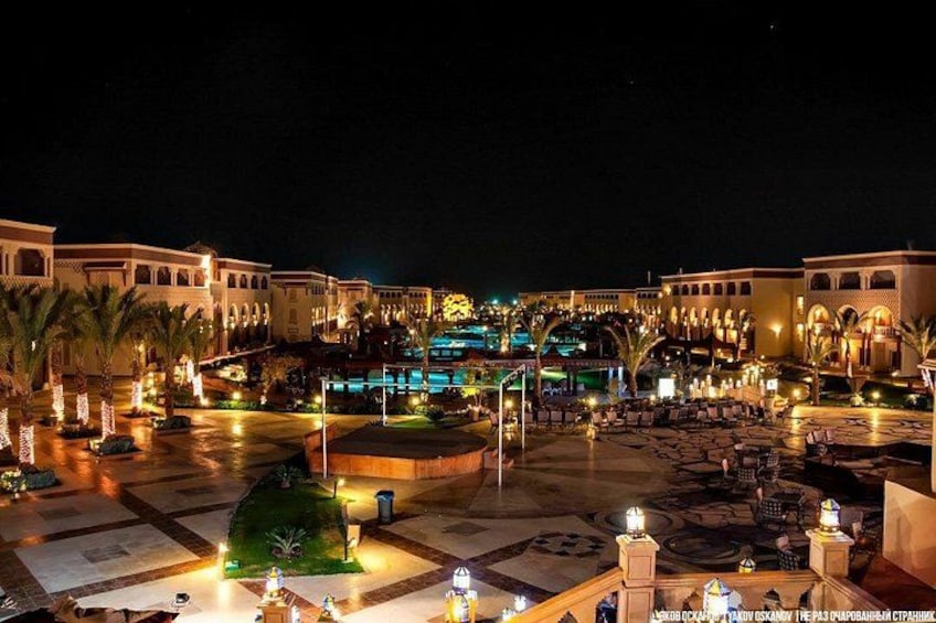 A well-lit resort courtyard at night with palm trees, a pool, and outdoor seating areas. 4