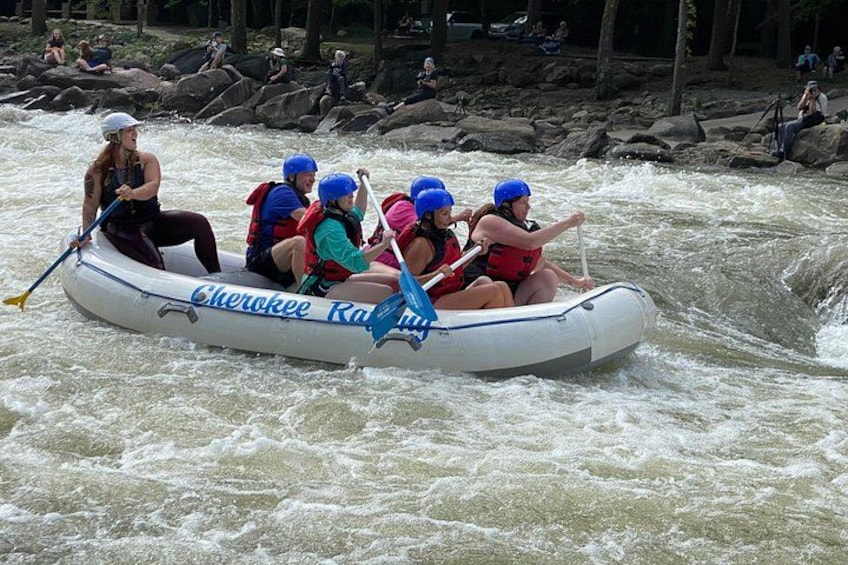 A group of people in a white inflatable raft with blue lettering, navigating a river with white water rapids. 3