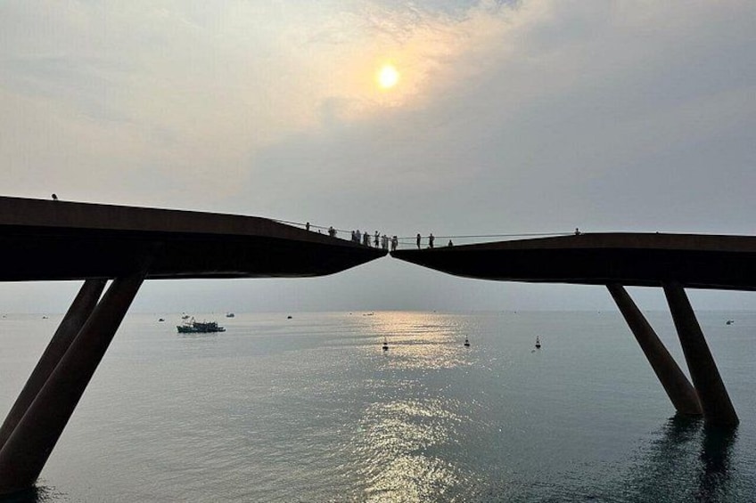A bridge with people walking on it, overlooking a calm body of water with boats and a setting sun. 1