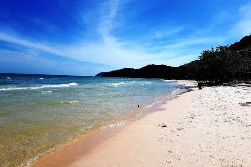 A beach with clear water, a sandy shore, and a forested hill in the background. 2