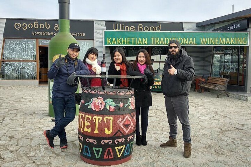 A group of people standing in front of a colorful barrel with text and a building with a sign that reads "Kakhetian Traditional Winemaking". 5