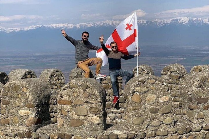 Two people standing on rocky terrain with a mountainous landscape in the background. 1
