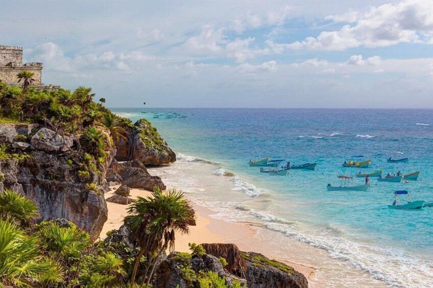 A beach with clear turquoise waters, boats, and a stone structure on a cliff. 5
