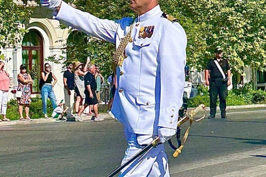 A person in a white military uniform with medals, holding a sword, standing on a street with spectators. 2