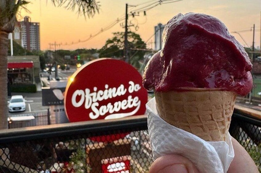 A hand holding an ice cream cone with a red topping, a sign for 'Oficina do Sorvete' in the background, and a cityscape with buildings and a road. 3