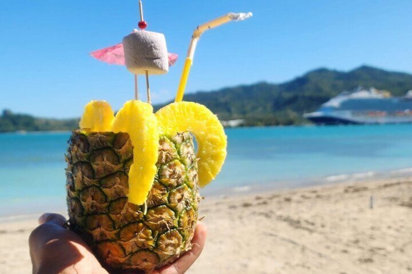 A hand holding a pineapple drink with a straw and a small umbrella on top, set against a beach and ocean backdrop. 2