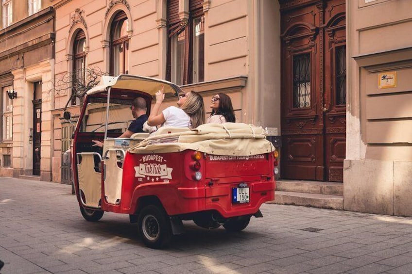 Een rode en beige Boedapest taxi met passagiers, geparkeerd op een geplaveid straatje voor een historisch gebouw. 4