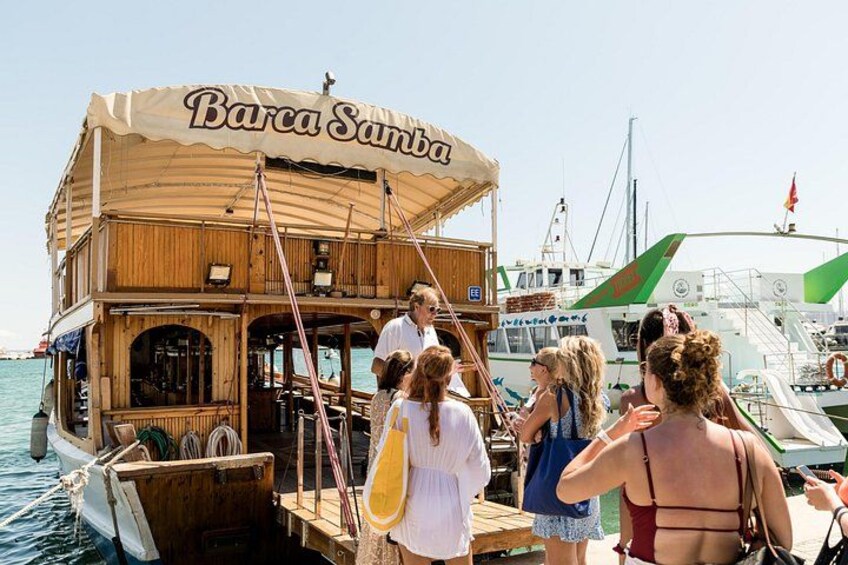 Een houten boot genaamd 'Barca Santa' ligt afgemeerd in een jachthaven, terwijl mensen aan boord gaan en van boord stappen. 3