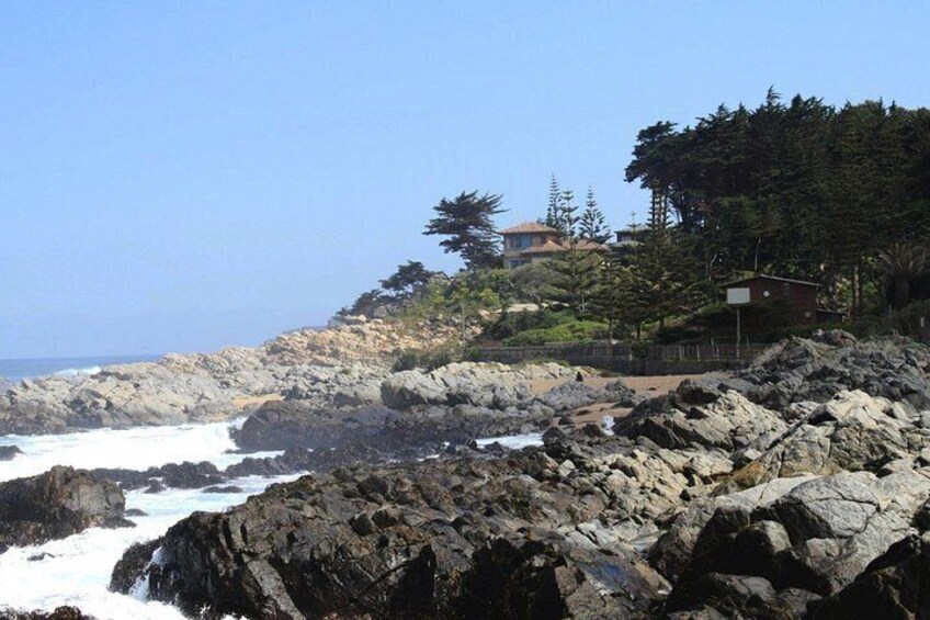 A rocky coastline with waves crashing against the rocks, a building on a hill, and dense trees. 5