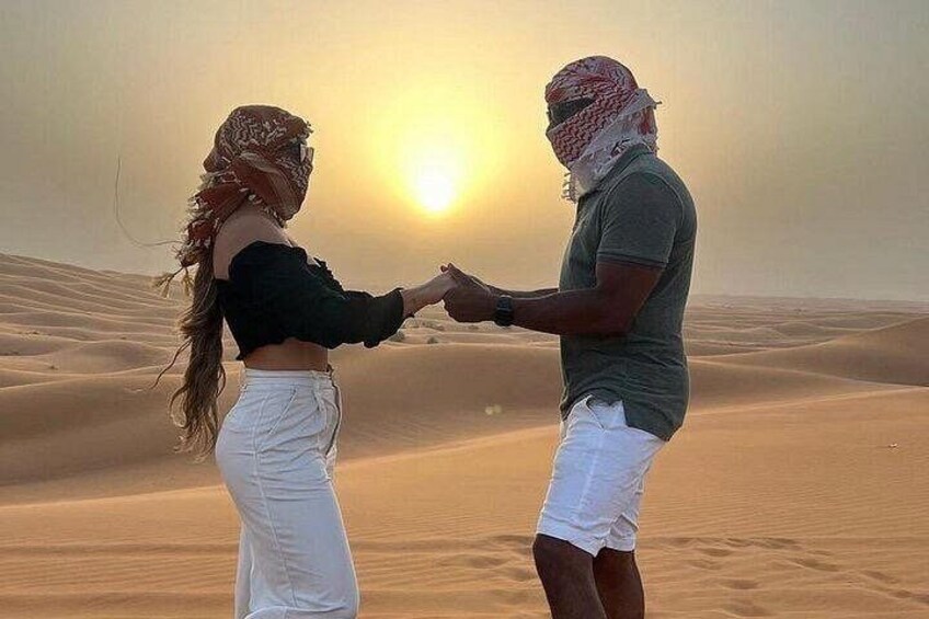 A couple holding hands in a desert landscape with sand dunes and a setting sun. 2