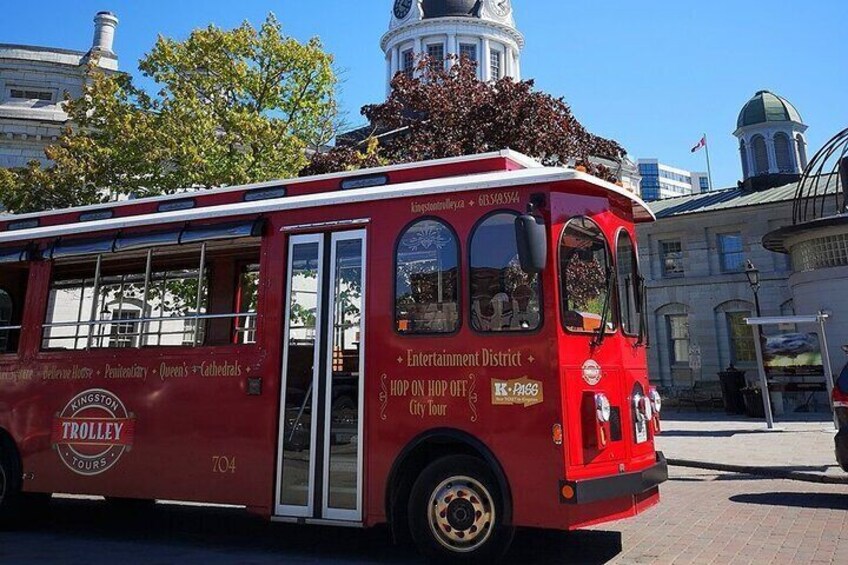 A red trolley with 'Entertainment District' and 'Hop On Hop Off City Tour' text, parked on a street with buildings and a clock tower in the background. 2
