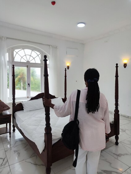 A person arranging a four-poster bed in a room with white walls, a large window, and marble flooring. 5