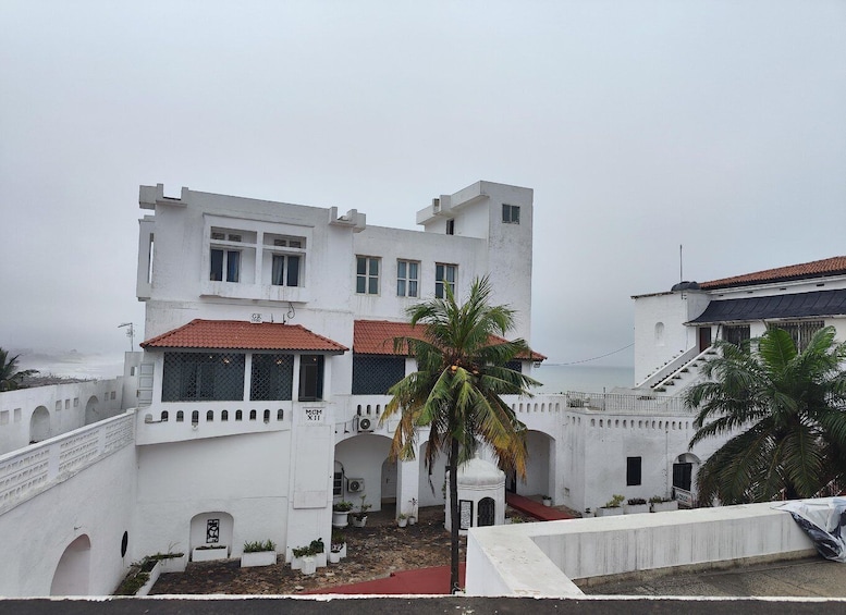 A white colonial-style building with a red-tiled roof, palm trees, and a coastal view. 1